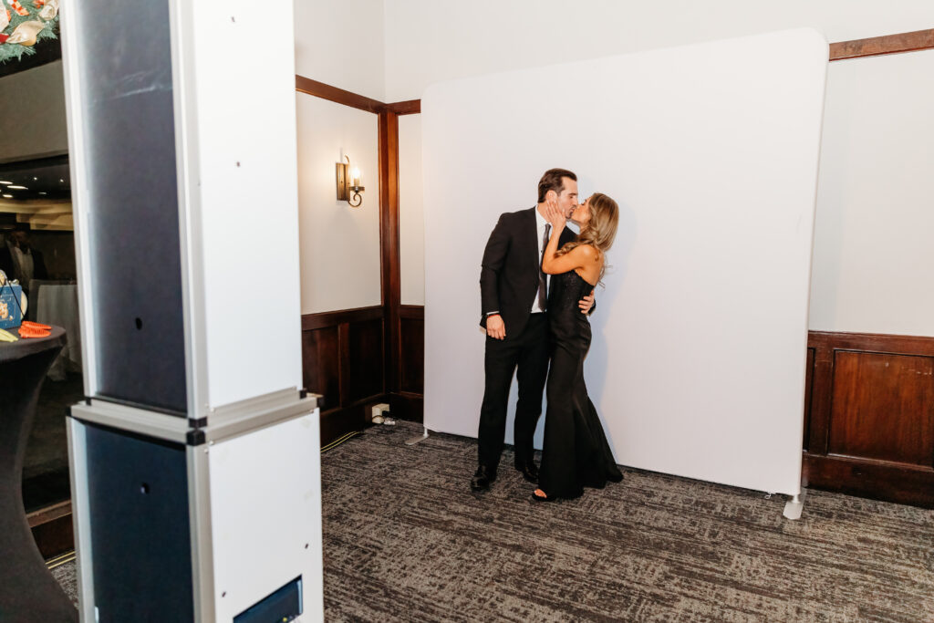 Two people dressed formally kiss in front of a white backdrop. The room features wooden paneling and a light fixture on the wall.