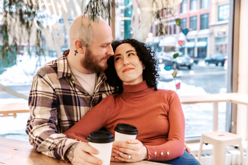 A person in a plaid shirt and another in an orange sweater share coffee and smiles in a cozy cafe with a snowy view.