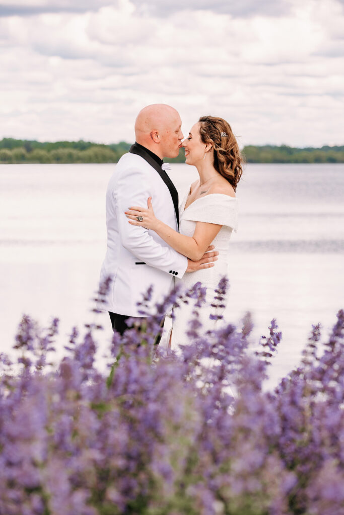Two people embracing by a lake, surrounded by purple flowers, with a cloudy sky and distant treeline in the background.