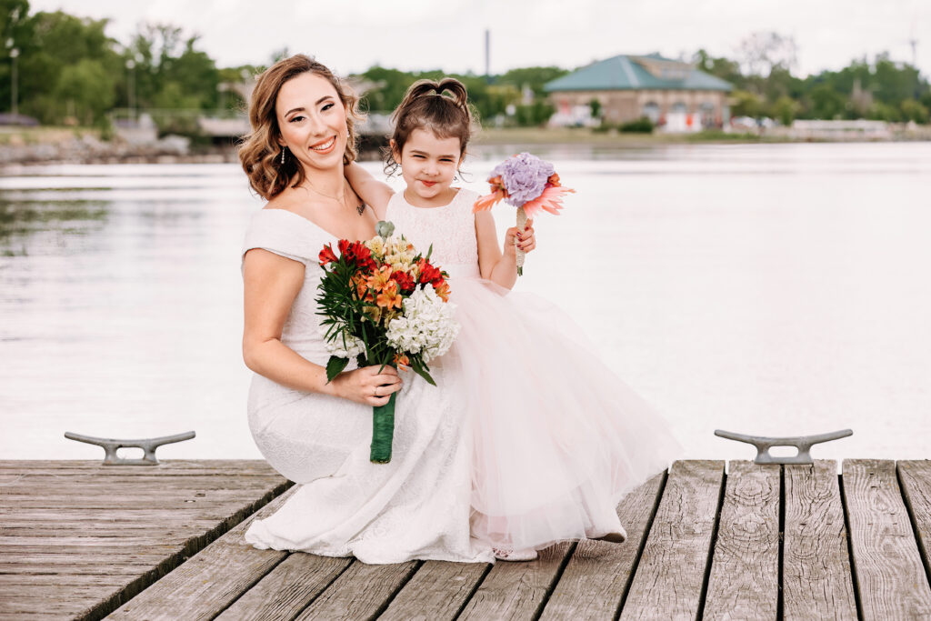 A person and child in white dresses hold flowers, smiling on a wooden dock by the water. Trees and a building in the background.
