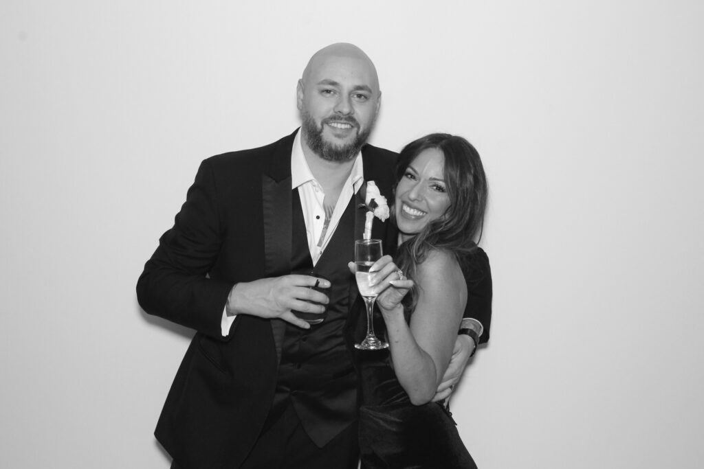 Two people in formal attire smiling, holding drinks, against a plain background. They appear to be at a celebration or event.