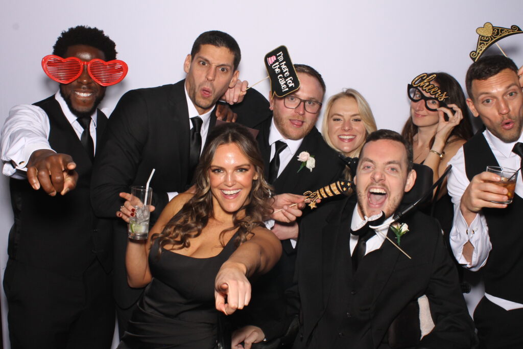 A group of eight people in formal attire celebrate, posing playfully with props in a photo booth setting. Everyone seems joyful and animated.