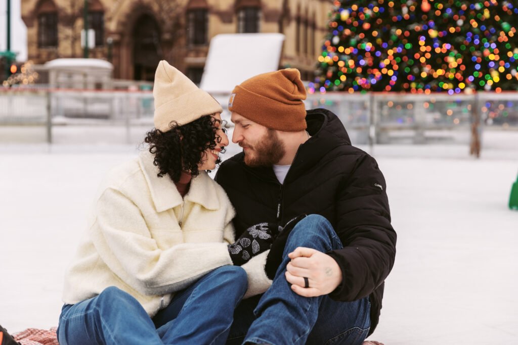 Two people sitting on an ice rink, smiling at each other, with a festive Christmas tree in the background, near historic building.
