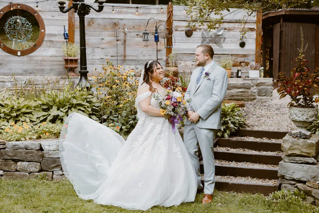 A person in a wedding dress and person in a suit stand in a garden with flowers, wooden fence, and stone steps.