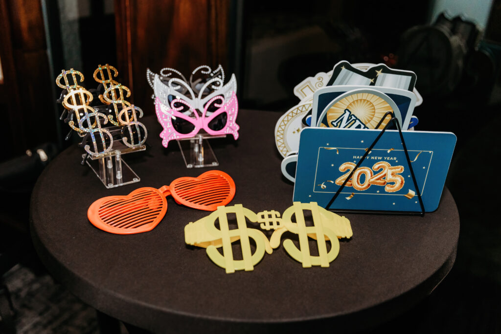 New Year 2025 party props including dollar sign glasses, heart-shaped eyewear, and festive cards, displayed on a round, dark table.