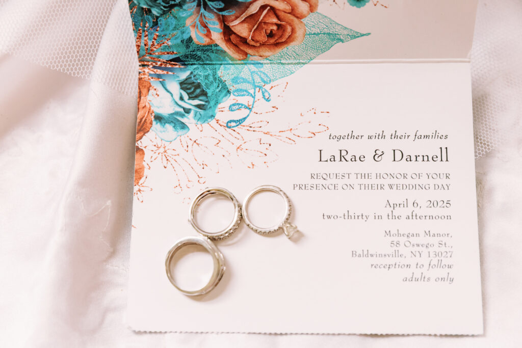 Wedding invitation with floral design and three rings displayed. Event at Mohegan Manor, Baldwinsville, NY, on April 6, 2025, at 2:30 PM.