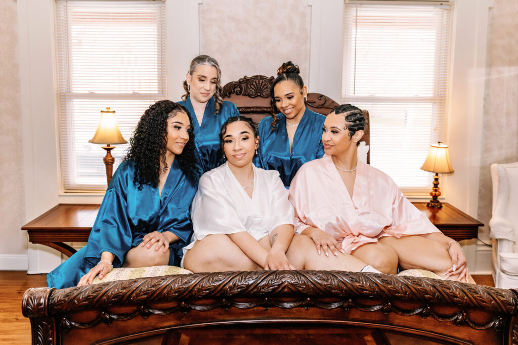 Five persons in colorful robes sit on a bed. The room features classic wooden furniture and soft lighting from table lamps.