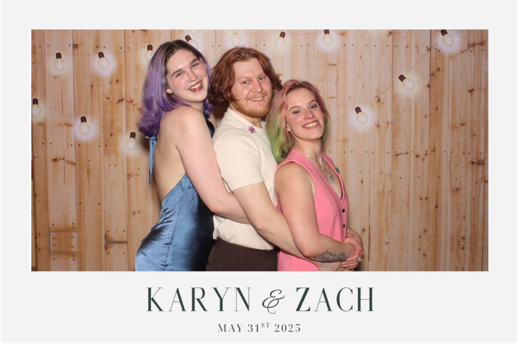 Three people smiling, posing in front of a wooden backdrop with string lights. Text reads "Karyn & Zach, May 31st, 2025."