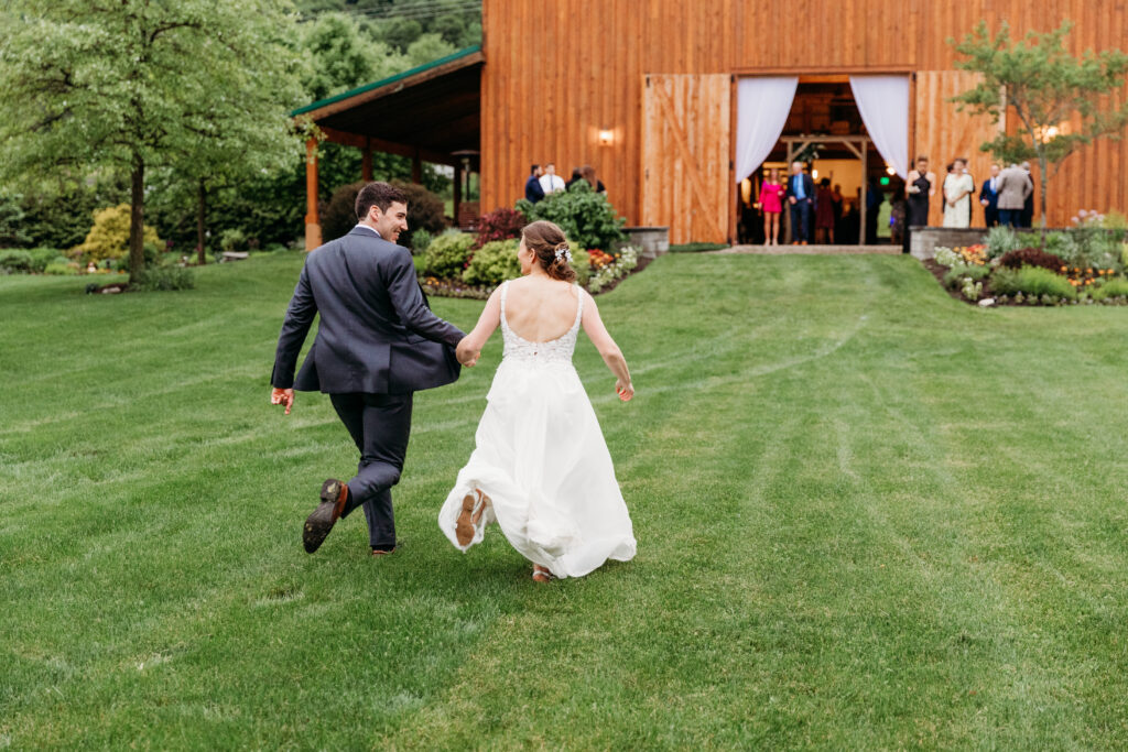 A person in formal attire and another in a white dress run toward a wooden barn, with people gathered outside on green grass.