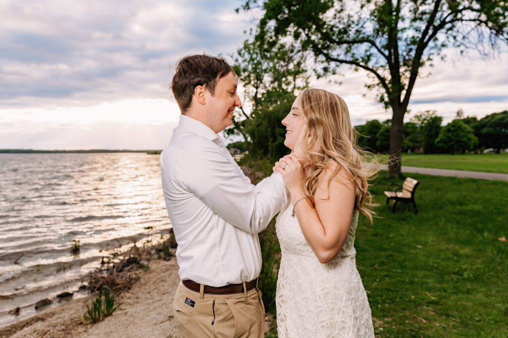 Two people smiling and holding hands by a lakeside park. The sun sets over the water, creating a serene, picturesque atmosphere.