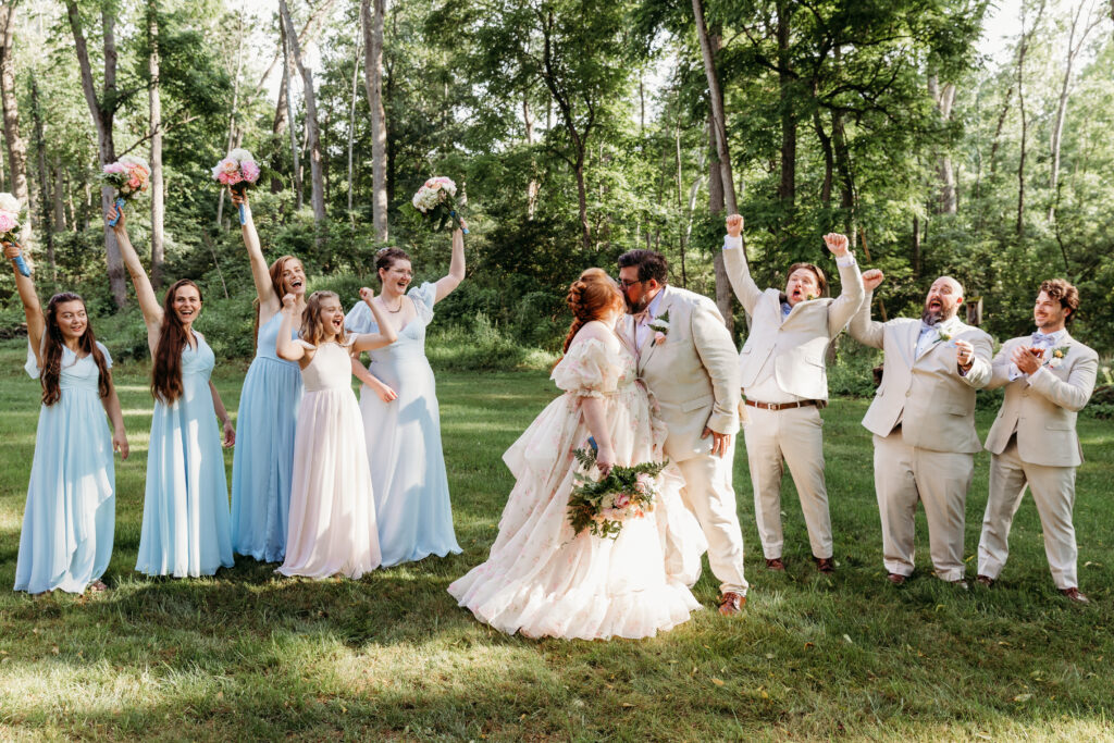 A wedding group celebrates outdoors, with a newlywed couple kissing surrounded by joyful persons in light blue and beige attire, holding bouquets.