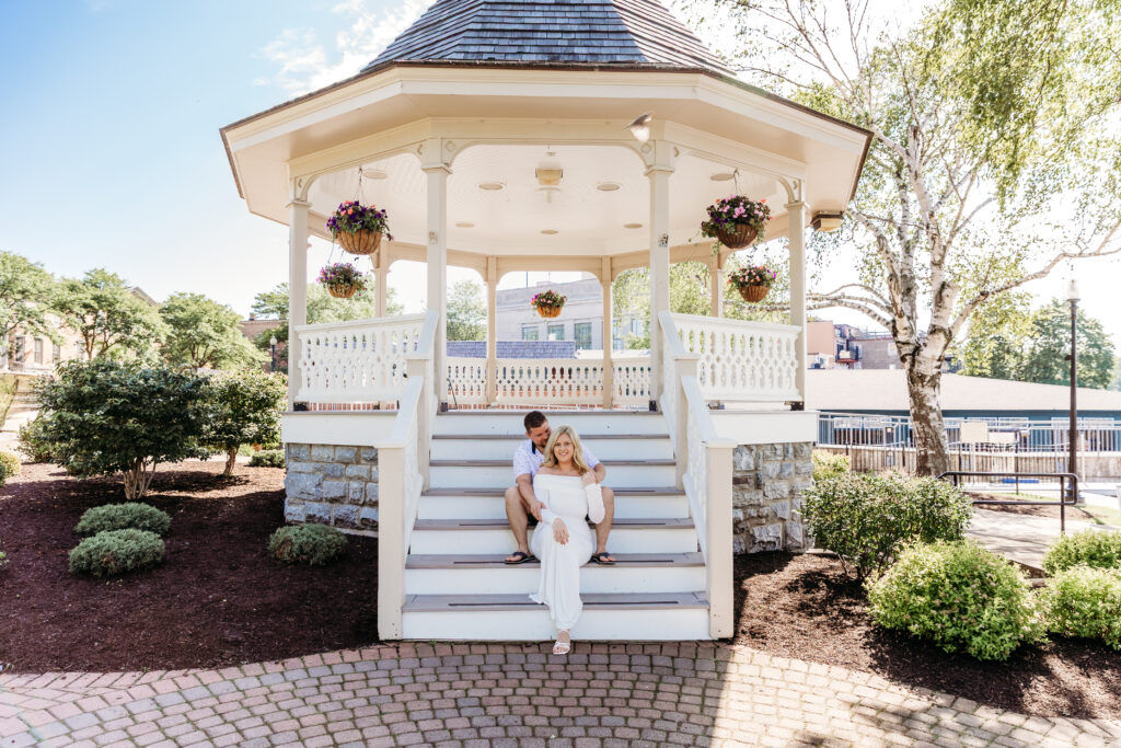A person in white sits with another on a gazebo staircase, surrounded by landscaped greenery and blooming flowers under a clear blue sky.
