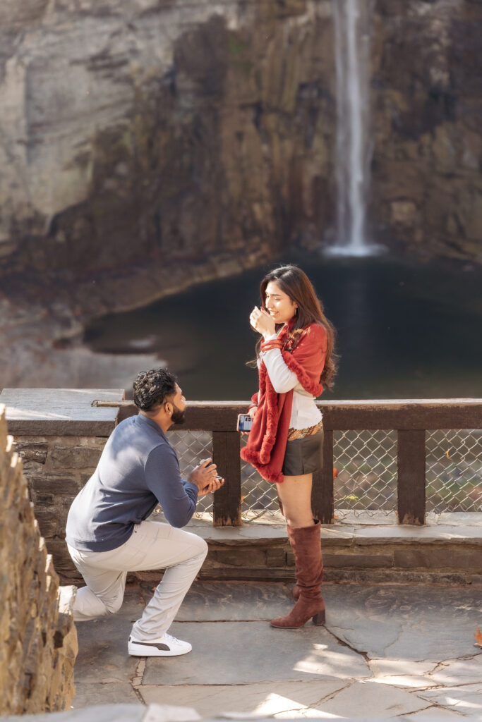 A person kneels, proposing to another person near a majestic waterfall, surrounded by rocky cliffs and a serene pool below.
