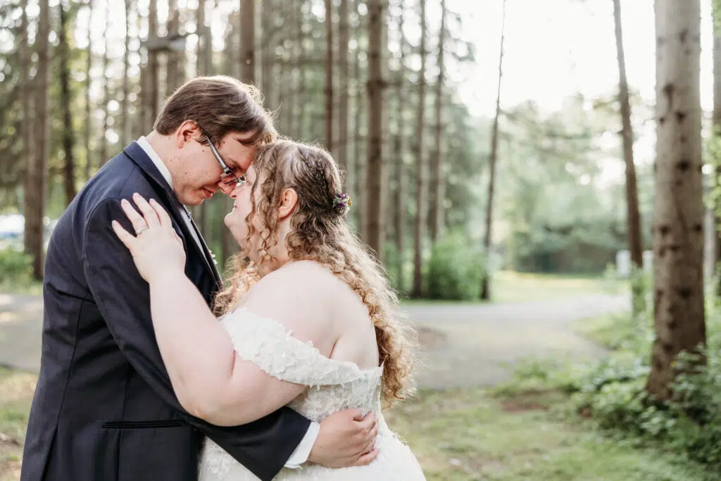 A bride in a white wedding dress and a groom in a dark suit kiss in a wooded outdoor setting.