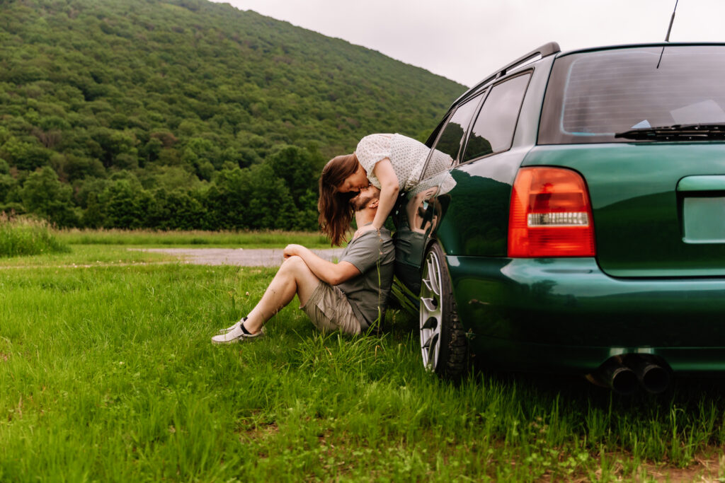A person sits beside a green car, interacting with another person in a grassy field near lush, green hills.