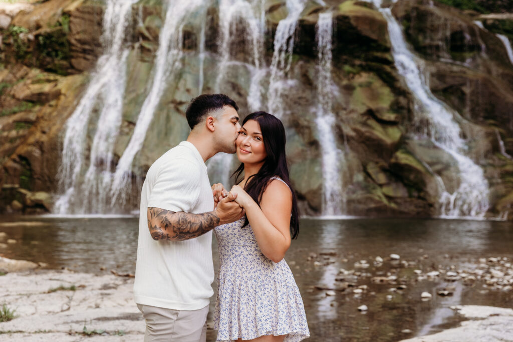Two people stand affectionately in front of a cascading waterfall, surrounded by natural greenery and rocky terrain, creating a serene outdoor scene.