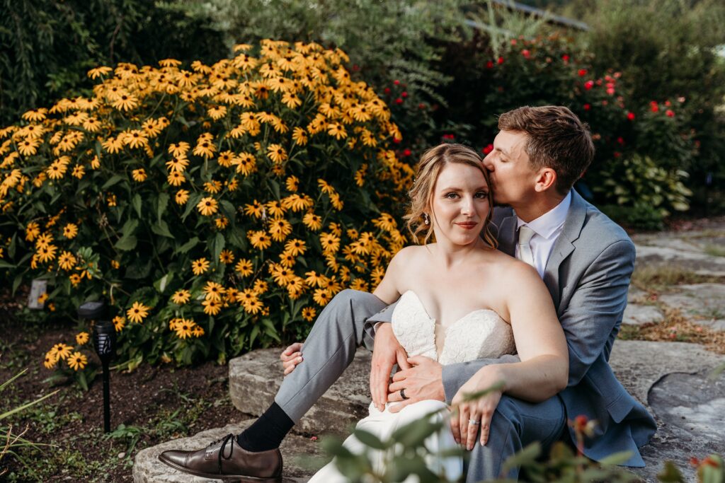 A bride and groom sit outdoors with a yellow-flowering bush in the background; the groom kisses the bride on the cheek as she smiles.