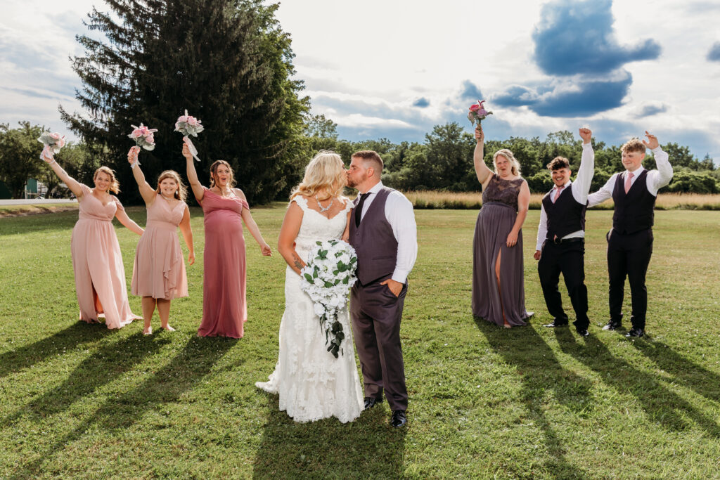 Bride and groom kiss in the center of a sunlit grassy field, flanked by bridesmaids in pink dresses with bouquets and groomsmen in vests cheering with arms raised.