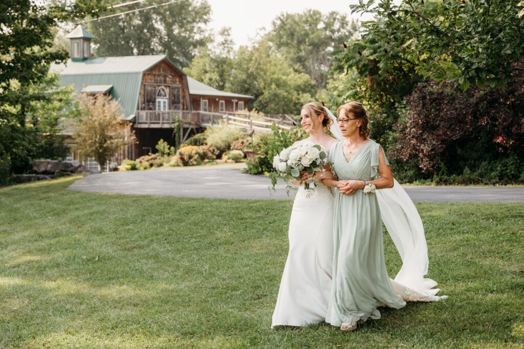 Two brides in pale dresses walk across a green lawn, holding bouquets, with a rustic house in the background.