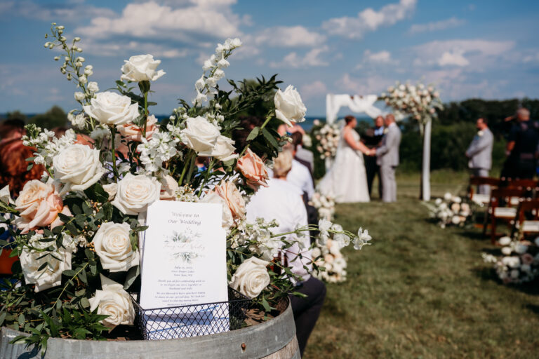 Outdoor wedding ceremony with three visible people. Focus on a flower arrangement and wedding program on a barrel. Clear sky in the background.