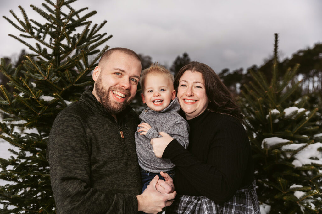 Three smiling people outdoors, surrounded by evergreen trees in a snowy setting. The two adults hold a child between them.