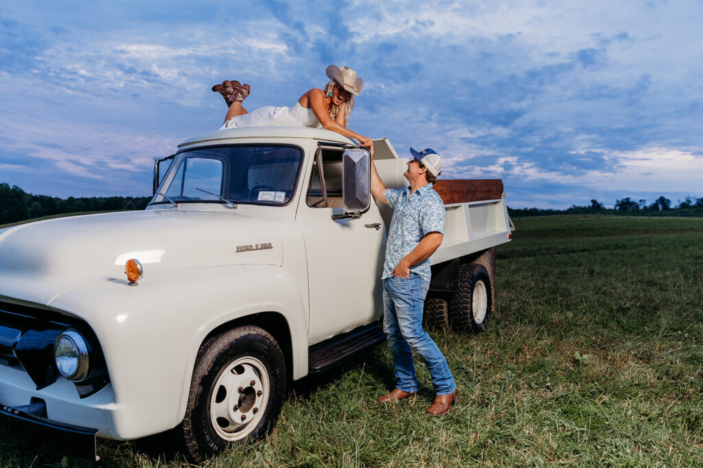 A person reclines atop a vintage Ford truck as another person stands beside it, both in a grassy field under a cloudy sky.