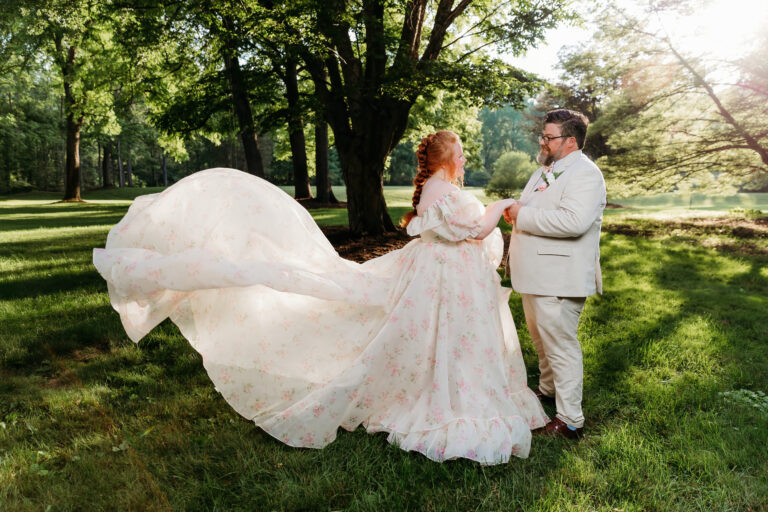 A bride and groom stand in a sunlit park, the bride in a flowing white wedding gown and the groom in a light suit.