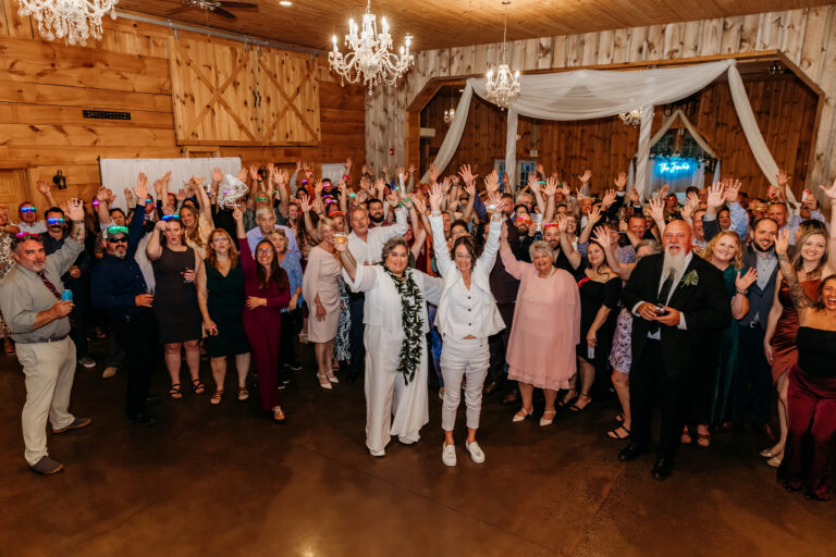 Guests at a rustic indoor wedding celebrate with the bride and groom in white at the center, raising their arms in joy.