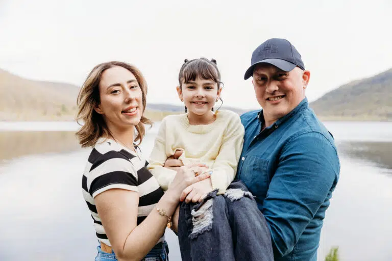 A smiling family of three posing outdoors by a lake with hills in the background.