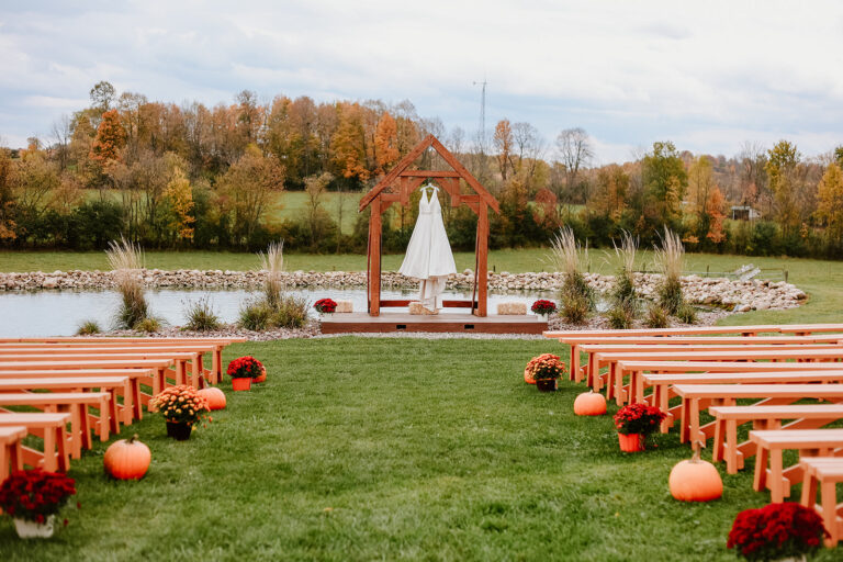 Autumn outdoor wedding ceremony setup with wooden benches along a grassy aisle, pumpkins and red flowers decorating the sides, and a white draped arch at the end by a lake.