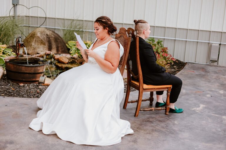 A bride in a white wedding dress sits on a chair reading a paper while a man in a black suit sits on another chair facing away in an outdoor courtyard with potted plants.