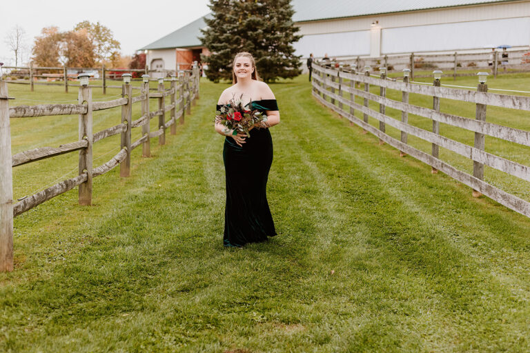 A woman in a long black dress holds a bouquet and walks along a grassy path between wooden fences on a farm.