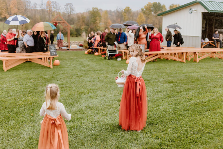 Two young girls in orange dresses stand on a grassy field as a crowd gathers around wooden picnic tables at an outdoor event.