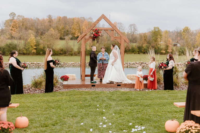 Outdoor autumn wedding by a lake with a wooden triangular arch and floral decorations, where the bride and groom stand under the arch as guests gather on a grassy lawn.