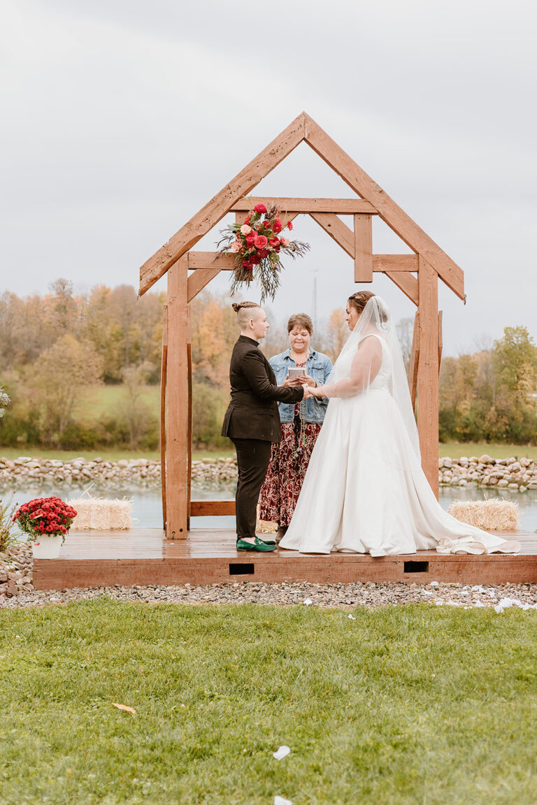 Bride and groom stand under a wooden arch with an officiant outdoors by a lake during their wedding.