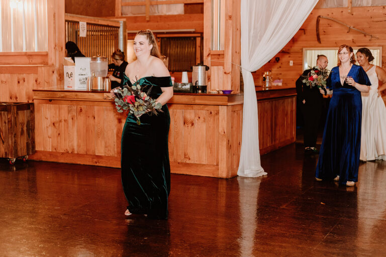 A woman in a black gown holding a bouquet walks across a wooden dance floor at a rustic venue, with other women in formal dresses in the background.
