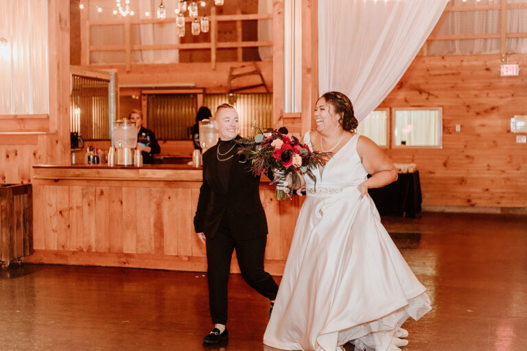 Bride in a white wedding gown and groom in a black suit dance together in a wooden reception hall, with the bride holding a bouquet.
