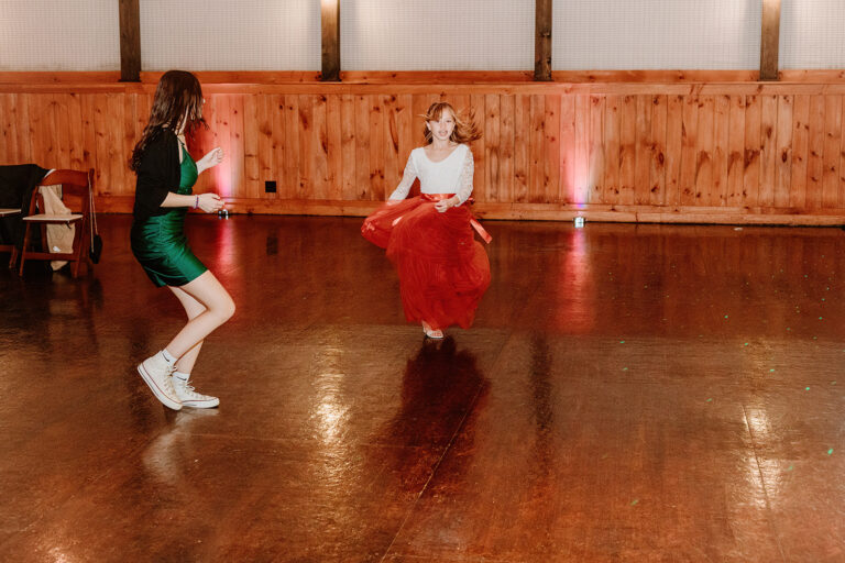 Two women dancing on a polished wooden floor in a wood-paneled hall, one in a green dress and white shoes and the other in a white top and red skirt.