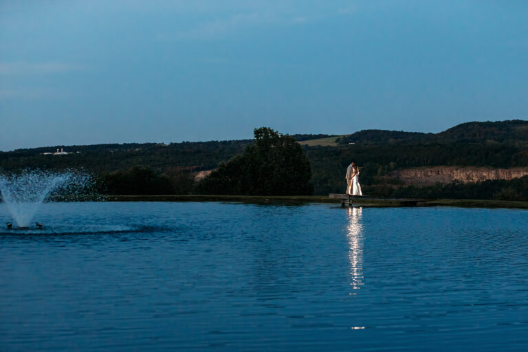 A calm lake with a fountain spray on the left and a white statue on the distant shore, with blue sky and tree-covered hills in the background.