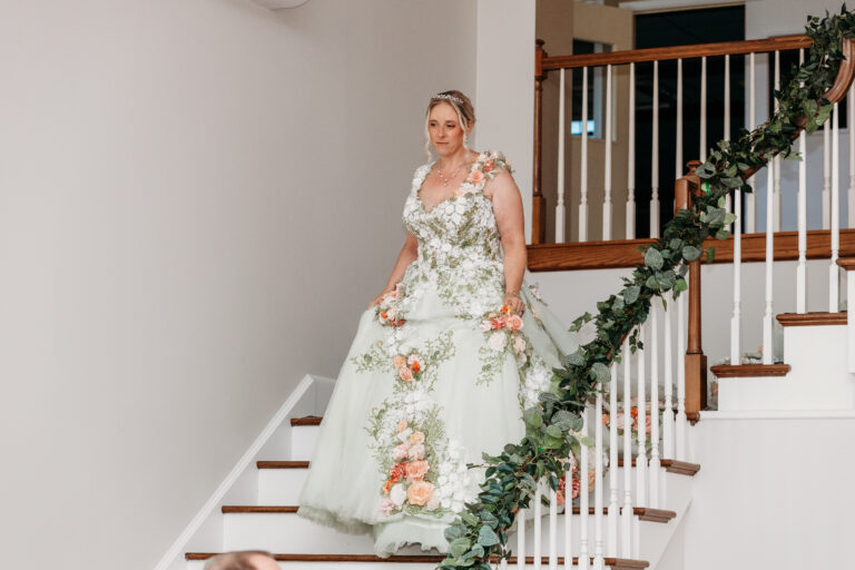 A woman in a pale green floral gown stands on a staircase adorned with a greenery garland along the railing.