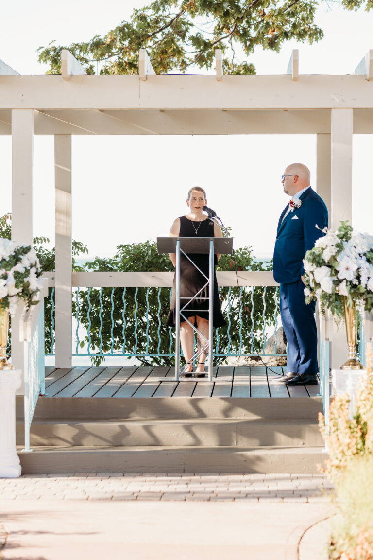 A man in a suit and a woman in a dress stand on a white gazebo platform with a lectern and microphone at an outdoor wedding ceremony, flanked by flower arrangements.