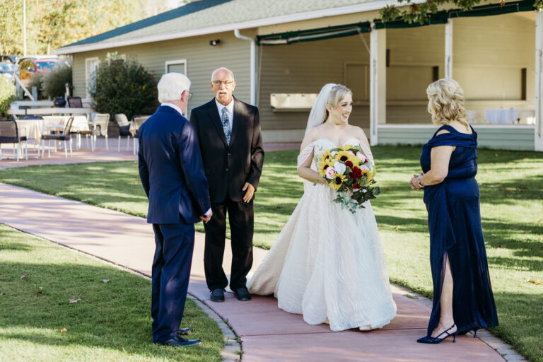Bride in a white wedding gown holding a bouquet stands on a brick path outdoors, greeting guests in formal attire.
