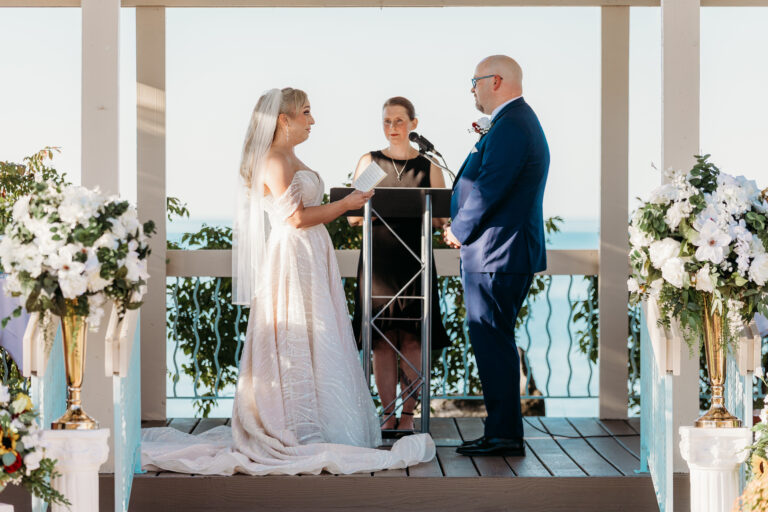 Bride and groom exchange vows with an officiant on an outdoor balcony, flanked by white floral arrangements and a seaside view.
