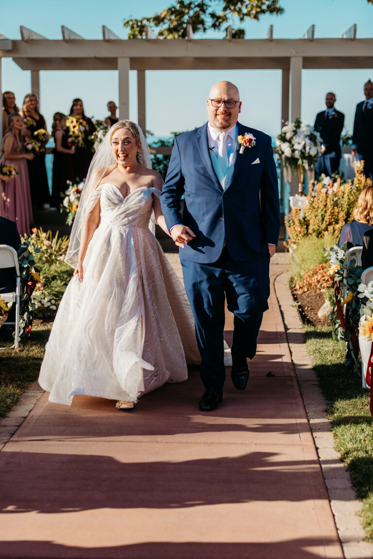 Bride in a pale pink wedding dress walks down an outdoor aisle arm-in-arm with an older man in a blue suit, as guests stand beside floral decorations.