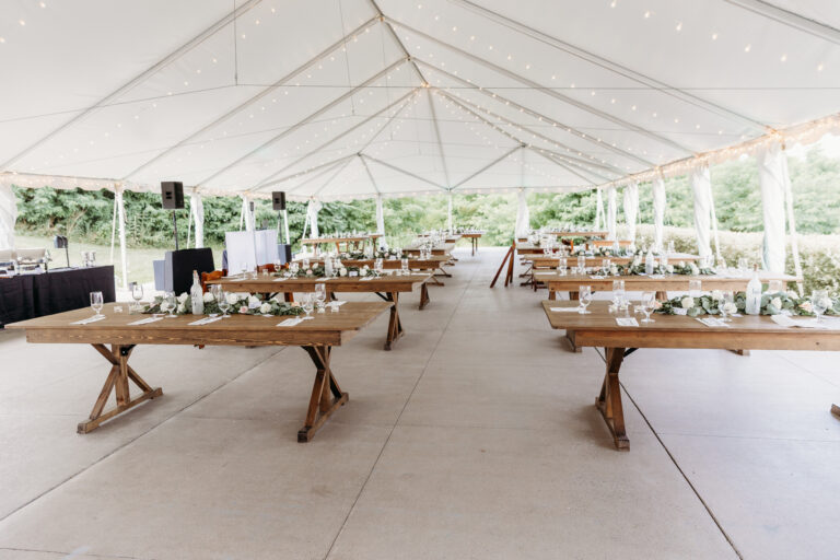 Long wooden tables with place settings and greenery inside a white event tent for an outdoor wedding.