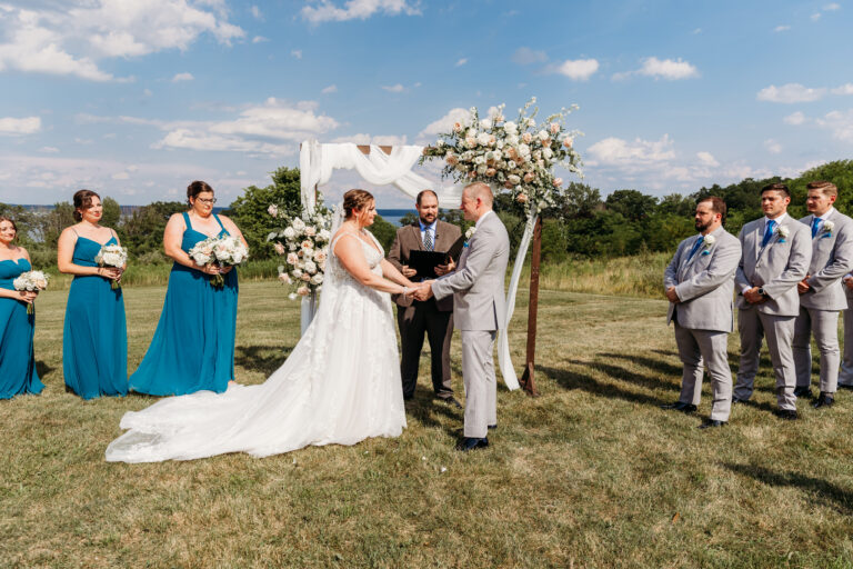 Outdoor wedding ceremony on a sunny grassy field with the bride in a white gown and the groom in a light gray suit exchanging vows under a floral arch, flanked by teal-dressed bridesmaids and gray-suited groomsmen.