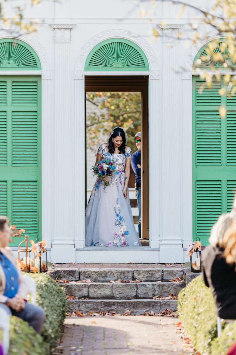 A bride in a floral wedding dress holding a bouquet stands in the doorway of a white building with green shutters, with guests seated outside.