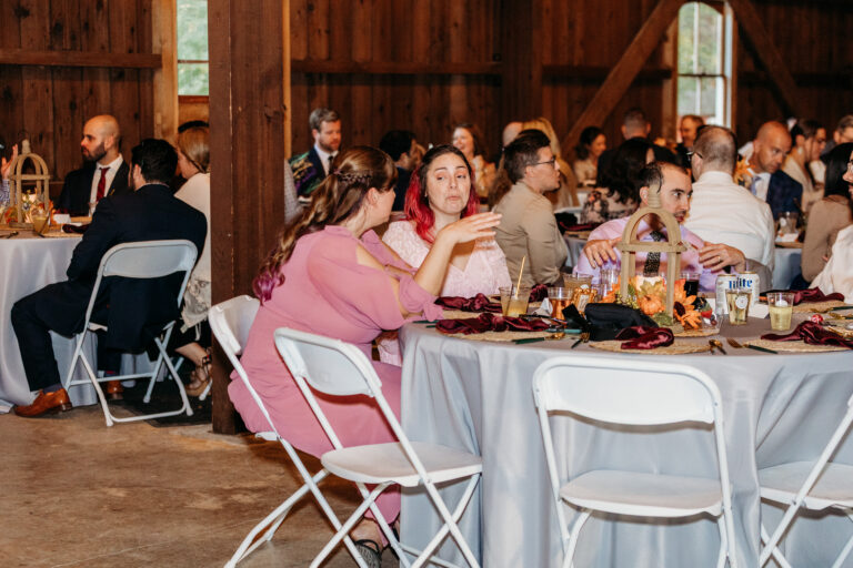 Guests sit at round tables with white chairs at a rustic barn wedding reception.