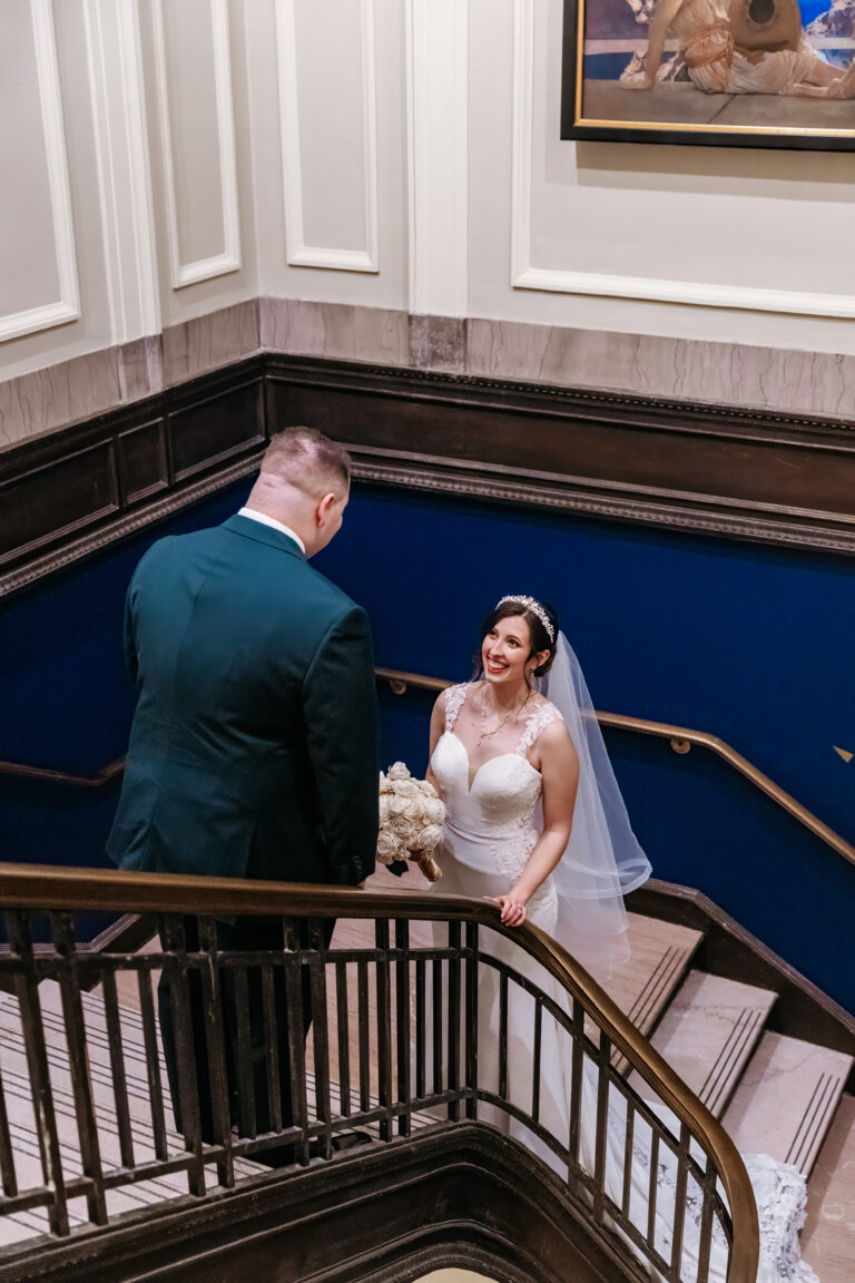 Bride in a white wedding dress and veil stands on a curved indoor staircase, holding a bouquet and smiling at her groom.