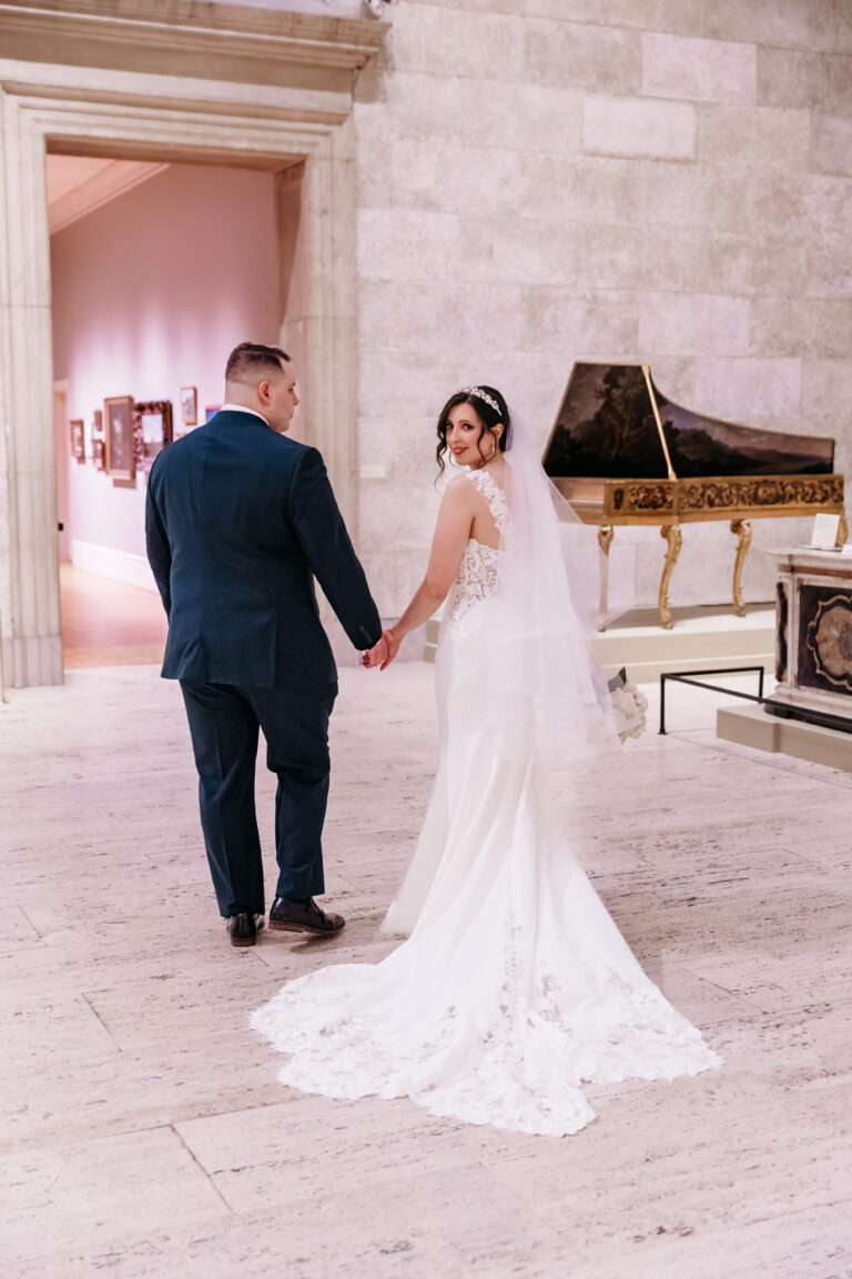 Bride in a white wedding gown with a long train and a groom in a dark suit walk hand in hand through a stone hallway toward a pink-lit doorway.